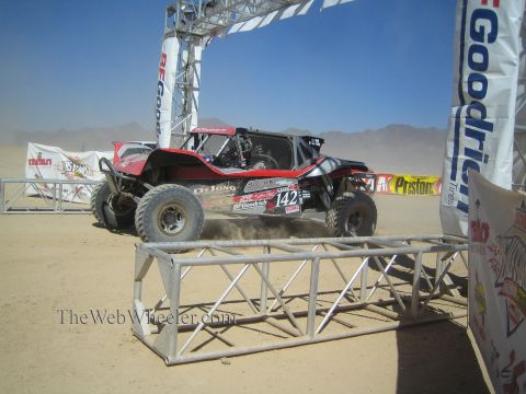 1990 Ford Bronco - red and black off-road race buggy numbered 142 with De Jong livery races beneath a BFGoodrich start arch in the desert.