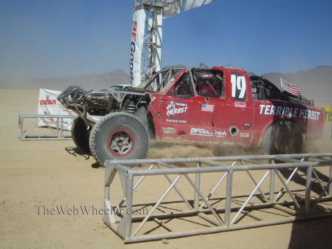 1990 Ford Bronco - red off-road race truck numbered 19 with Terrible Herbst livery races from a desert start line under a banner arch.