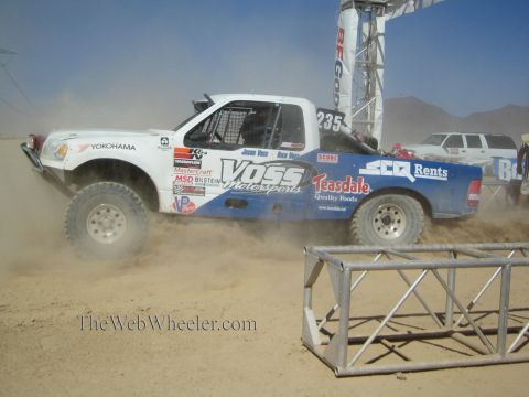1990 Ford Bronco - white and blue off-road race truck numbered 235 with Voss Motorsports livery races across a dusty desert course.