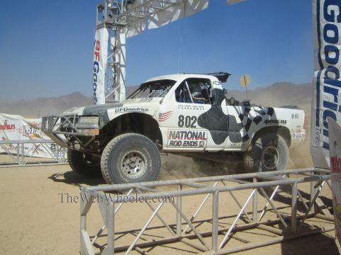 1990 Ford Bronco - white off-road race truck numbered 802 with cow-print graphics and National Rod Ends livery races at a desert start arch.