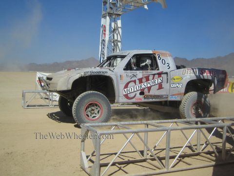 1990 Ford Bronco - white off-road race truck numbered 803 with Owl Motorsports livery races over a berm at a desert start arch in dust.