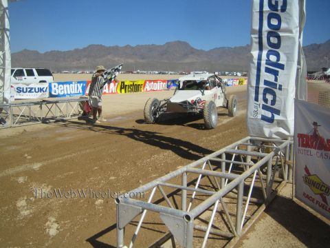 1990 Ford Bronco - white off-road race buggy approaches a Goodrich-banner finish arch while a flagman signals on a sandy desert course.