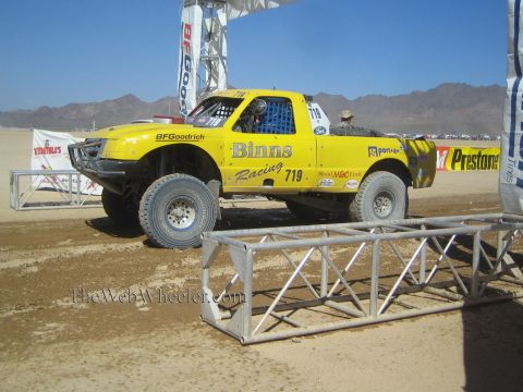 1990 Ford Bronco - yellow Binns Racing number 719 off-road race truck sits on a desert course beside metal barriers under a clear blue sky.