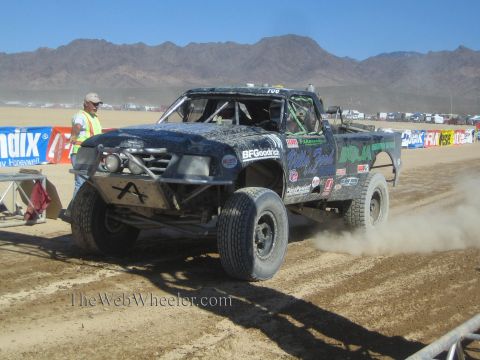 1990 Ford Bronco - dirt-covered dark blue off-road race truck drives along a dusty desert track as a man in a safety vest watches.