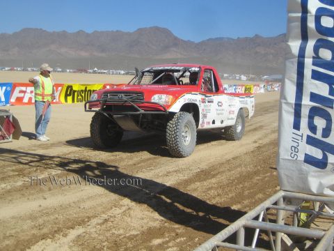 1990 Ford Bronco - red and white off-road race truck sits on a churned dirt course as a man in a safety vest stands nearby.