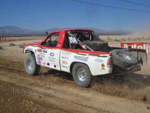1990 Ford Bronco - white number 204 off-road race truck carrying spare tires kicks up dust along a desert race trail.