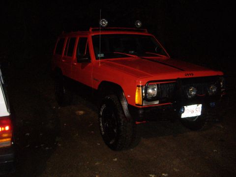 1990 Jeep Cherokee - A red 1990 Jeep Cherokee is seen from a three-quarter front view at night, with another vehicle partially visible on the left.