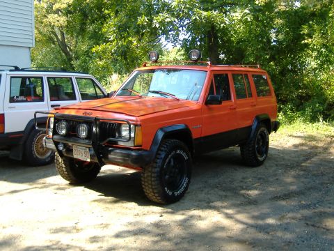 1990 Jeep Cherokee - An orange 1990 Jeep Cherokee is parked outdoors next to a white Jeep, a roof rack and a front bull bar.