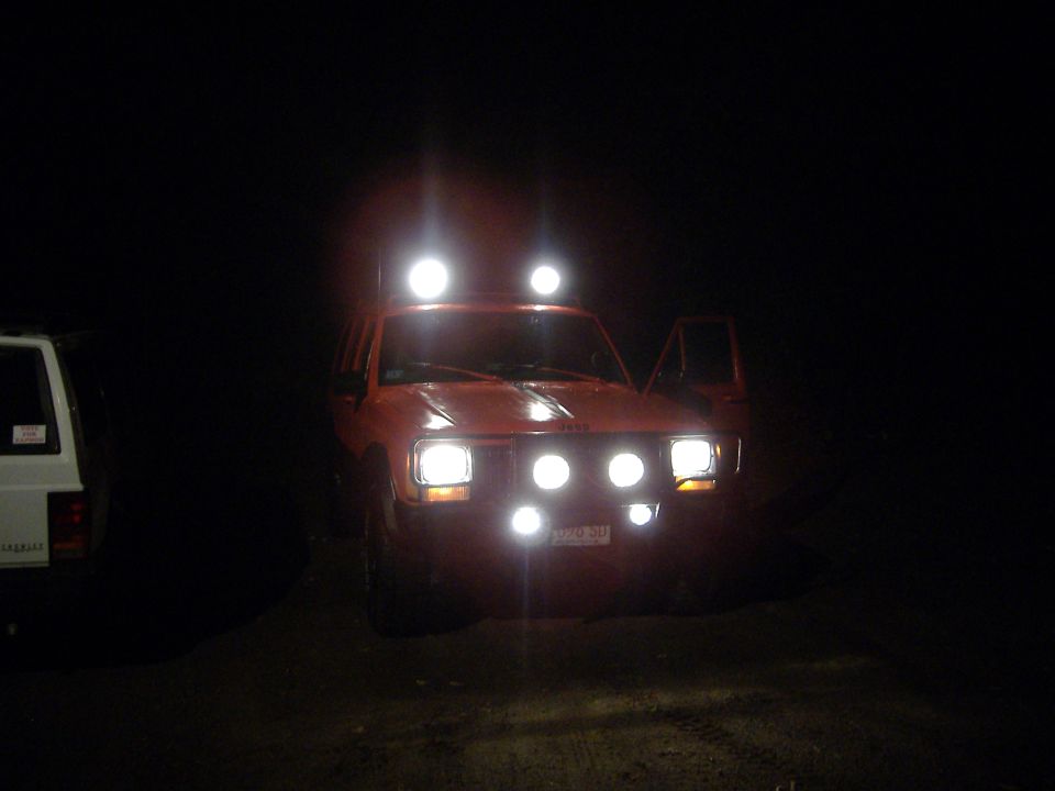 Orange 1990 Jeep Cherokee with auxiliary lights on roof and bumper, driver's door open. White SUV parked to the left