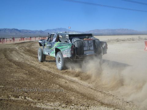 1990 Ford Bronco - black and green off-road race truck speeds along a dusty desert course, throwing up a cloud of dust.