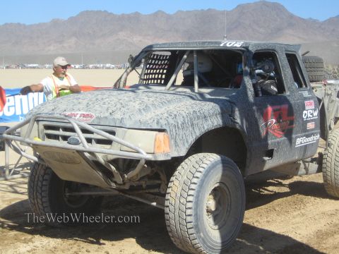 1990 Ford Bronco - gray off-road race truck coated in dust sits on a desert course while a man in a safety vest stands at left.