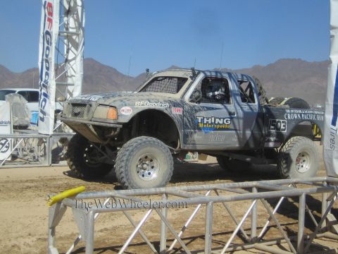 1990 Ford Bronco - gray number 1503 off-road race truck lettered Thing Motorsports sits on a dirt course behind metal barriers.
