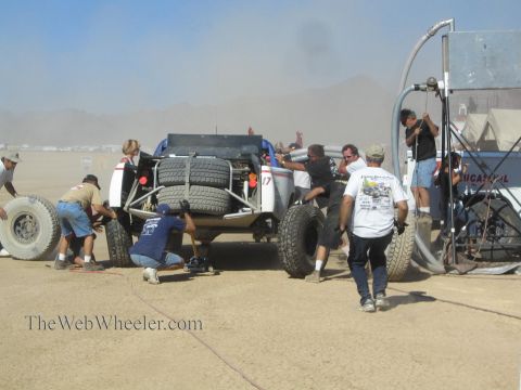 1990 Ford Bronco - pit crew works around an off-road race truck loaded with spare tires in a dusty desert refueling pit.