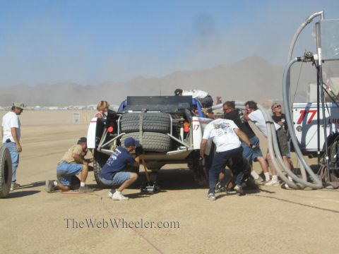 1990 Ford Bronco - Pit crew members kneel and work at the rear of an off-road race truck carrying spare tires in a dusty desert pit.