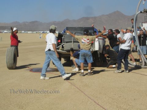 1990 Ford Bronco - crew member raises both arms beside an off-road race truck while others work around it in a dusty desert pit.