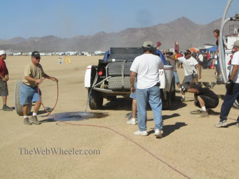 1990 Ford Bronco - crew member sprays water from a hose near a number 17 off-road race truck surrounded by pit crew in the desert.