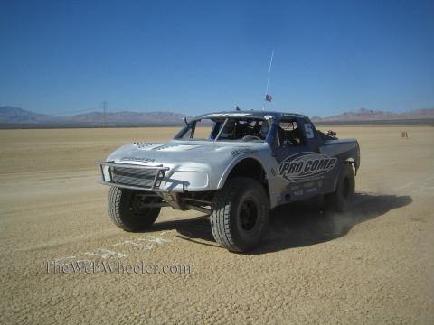 1990 Ford Bronco - silver Pro Comp number 5 off-road race truck drives across a flat desert course under a clear blue sky.