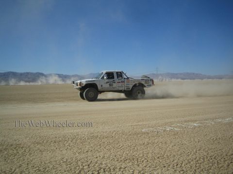 1990 Ford Bronco - white off-road race truck speeds across an open desert course, leaving a long trail of dust behind it.