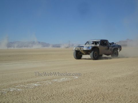 1990 Ford Bronco - off-road race truck races across a hazy open desert flat, kicking up dust under distant mountains.