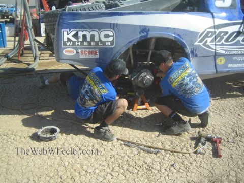 1990 Ford Bronco - Two crew members in blue shirts kneel to service the rear wheel hub of a blue off-road race truck with KMC Wheels decals.
