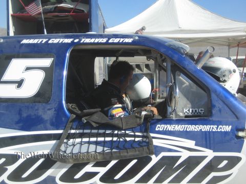 1990 Ford Bronco - driver sits inside the cab of a blue number 5 Coyne Motorsports off-road race truck under a pit canopy.