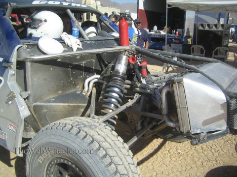 1990 Ford Bronco - exposed front suspension, coilover shock, and tube chassis of an off-road race truck are visible in a shaded pit.