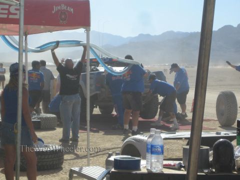 1990 Ford Bronco - crew works under a Jim Beam canopy servicing an off-road race truck amid spare tires in a dusty desert pit.