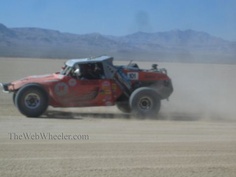 1990 Ford Bronco - orange tube-frame off-road race buggy carrying number 101 races across a dusty desert lakebed with mountains behind.