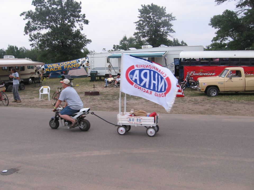 Man on mini bike pulls wagon with NHRA flag; RVs and pickup truck in background