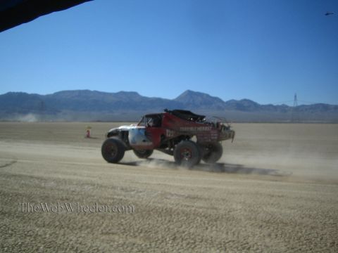 1990 Ford Bronco - off-road race truck throws up dust as it speeds across a wide desert flat under a clear blue sky.
