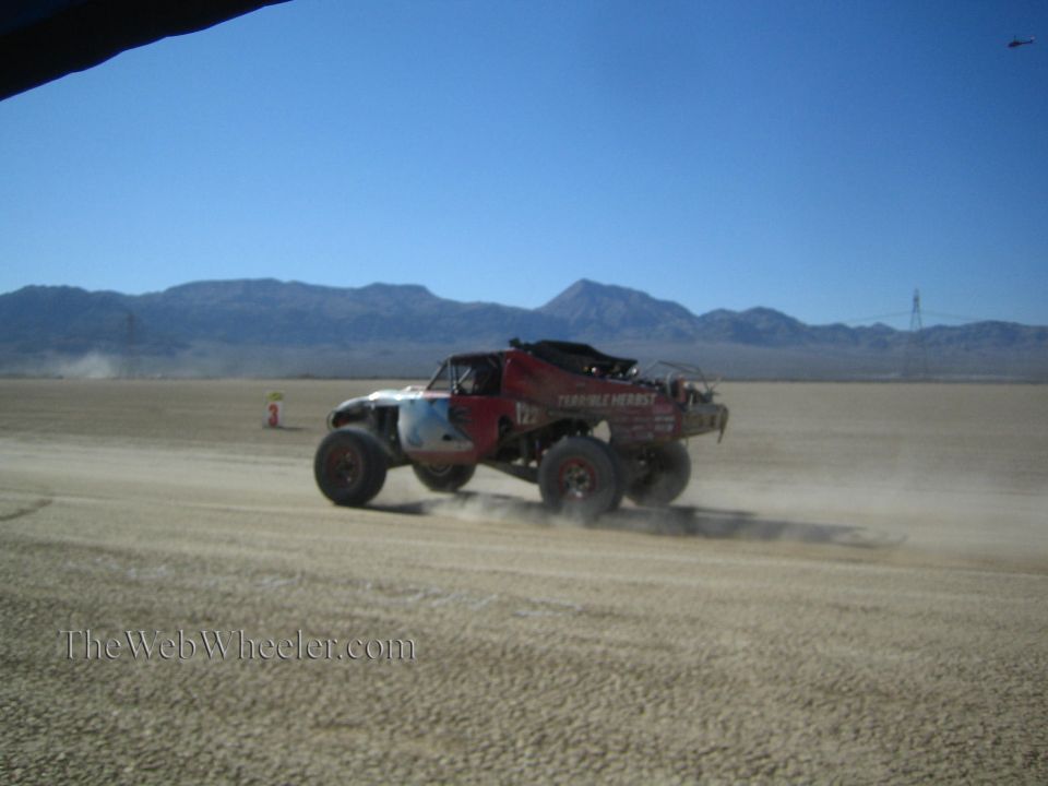 Off-road race truck speeding across open desert flat with power lines and mountains