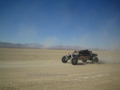 1990 Ford Bronco - long-travel off-road race buggy speeds across a hazy desert lakebed with a power-line tower on the horizon.