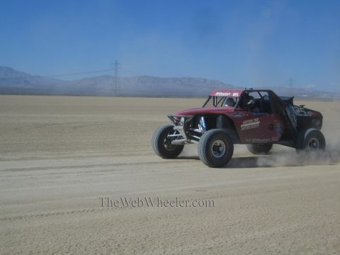 1990 Ford Bronco - dark red tube-frame off-road race buggy races across a desert flat, trailing dust beneath a clear sky.