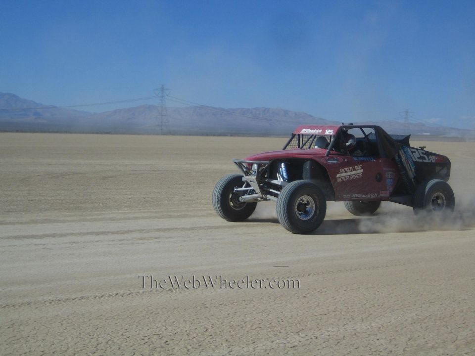 Dark red off-road race buggy speeding across desert flat trailing dust