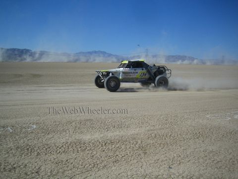 1990 Ford Bronco - white and green off-road race buggy marked 134 kicks up dust racing across a cracked desert lakebed.