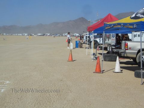 1990 Ford Bronco - Canopy pit tents and a pickup line a desert race staging area marked with traffic cones, mountains in the distance.