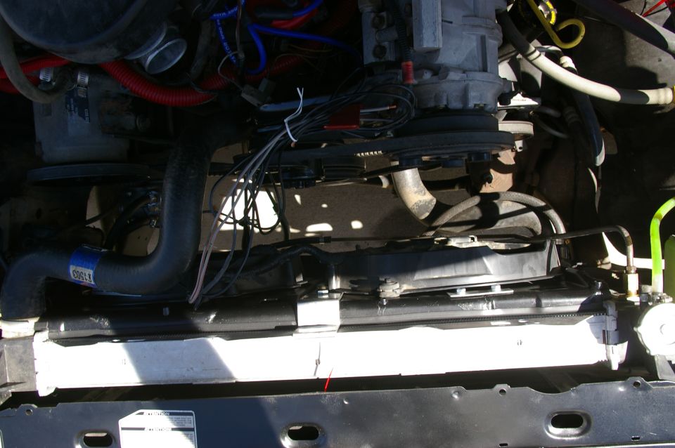 Engine bay showing radiator, fan, hoses, and wiring. A blue label on a hose is visible
