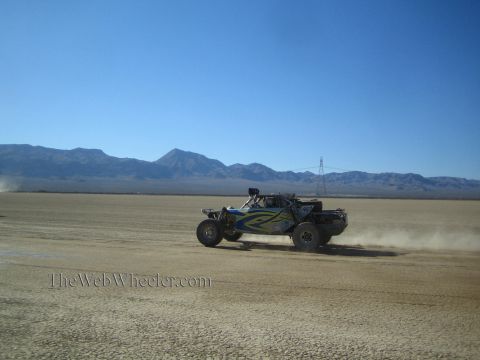 1990 Ford Bronco - blue and green off-road race buggy speeds across an open desert flat with rugged mountains on the horizon.