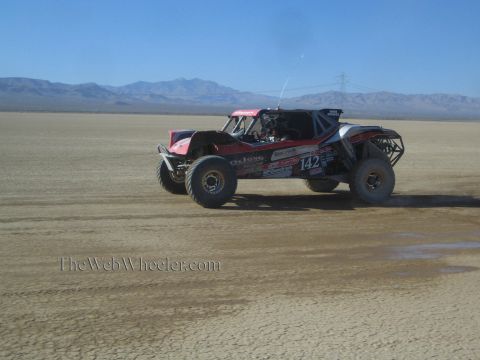 1990 Ford Bronco - black and red off-road race buggy carrying number 142 races across a desert lakebed with distant mountains.