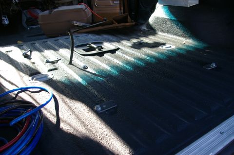 1986 Ford Bronco - The interior of a truck bed shows a textured dark liner with several tie-down points and a bundle of blue and red cables.