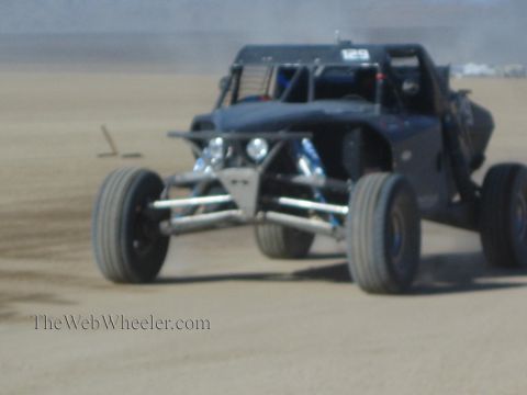 1990 Ford Bronco - dark long-travel off-road race buggy marked 129 charges toward the camera across a dusty desert course.