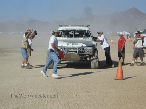1990 Ford Bronco - crew pushes a white off-road race truck through a dusty desert pit while photographers and onlookers gather around cones.