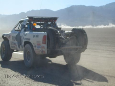 1990 Ford Bronco - white off-road race truck speeds away across a dusty desert flat with mountains rising in the haze behind.