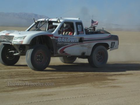 1990 Ford Bronco - white Baldwin Motorsports off-road race truck number 97, flying a small flag, races across the dusty desert.