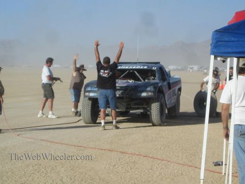 1990 Ford Bronco - man raises both arms beside a dark off-road race truck as crew and onlookers gather at a dusty desert pit.