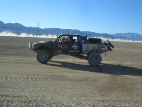 1990 Ford Bronco - black off-road race truck carrying number 702 races across a hazy desert lakebed with mountains behind.
