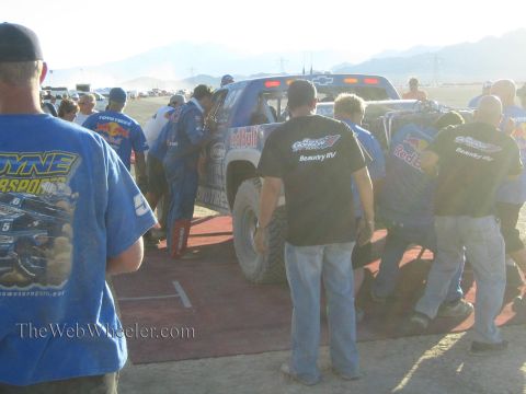 1990 Ford Bronco - crowd of crew in team shirts surrounds a race truck parked on a red pit mat in a hazy, dusty desert pit.