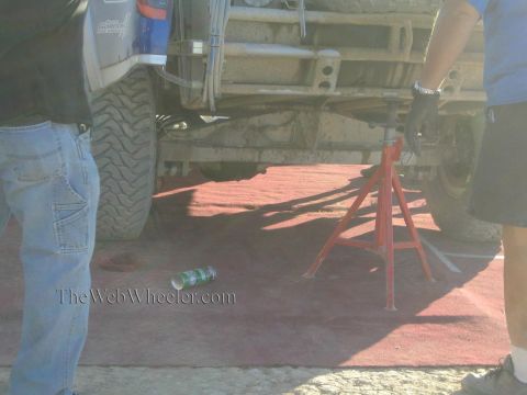 1990 Ford Bronco - race truck sits on a red jack stand showing its leaf springs and tires while crew stand on a red pit mat.