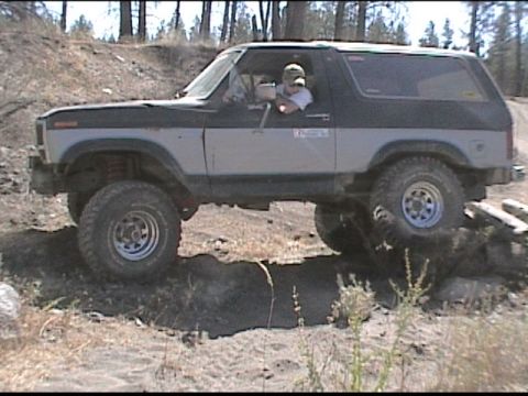 1985 Ford Bronco - A two-tone Ford Bronco navigates a challenging off-road course, with its tires on uneven ground.