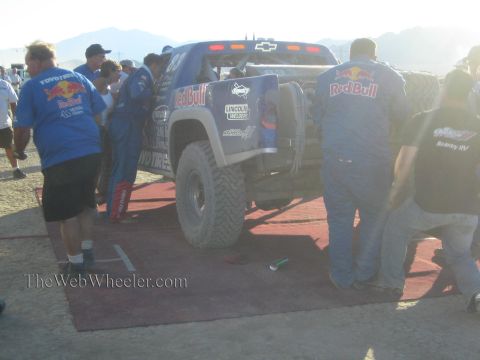 1990 Ford Bronco - pit crew in Red Bull and Toyo Tires shirts gathers around the rear of a blue Chevrolet trophy truck on a red mat in hazy desert light.